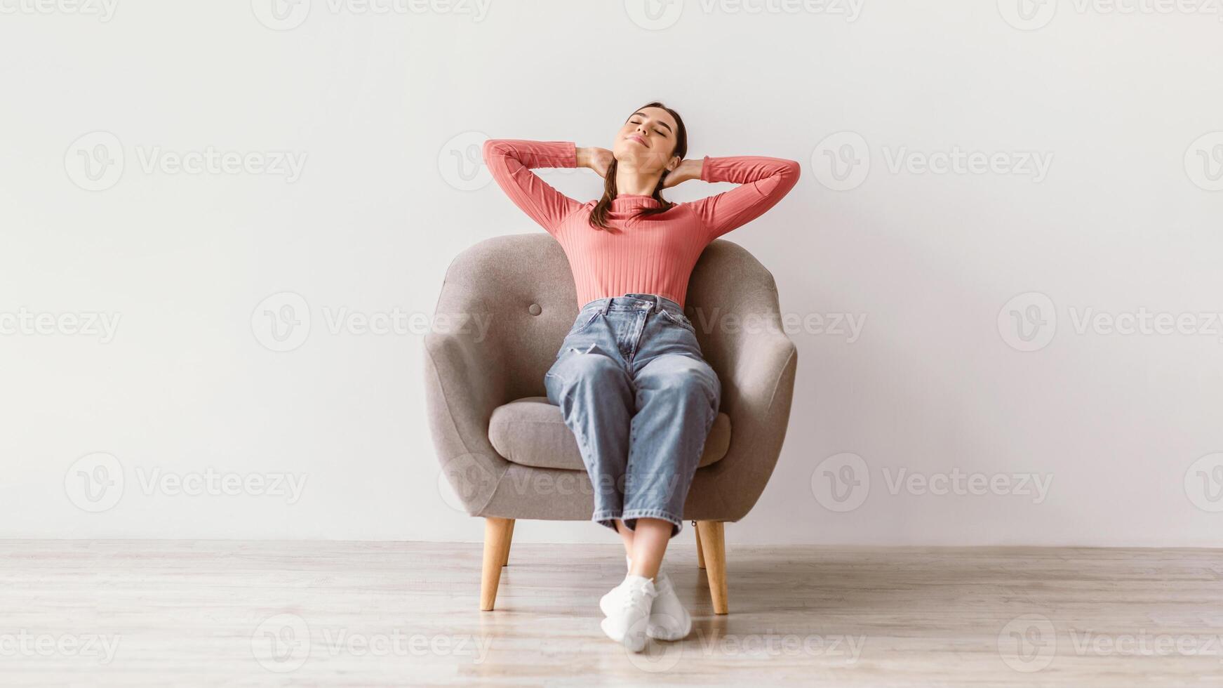 A young brunette woman enjoys a moment of relaxation while sitting in a cozy armchair. She stretches her arms behind her head in a stylish, minimalistic indoor setting, embracing a peaceful lifestyle. photo