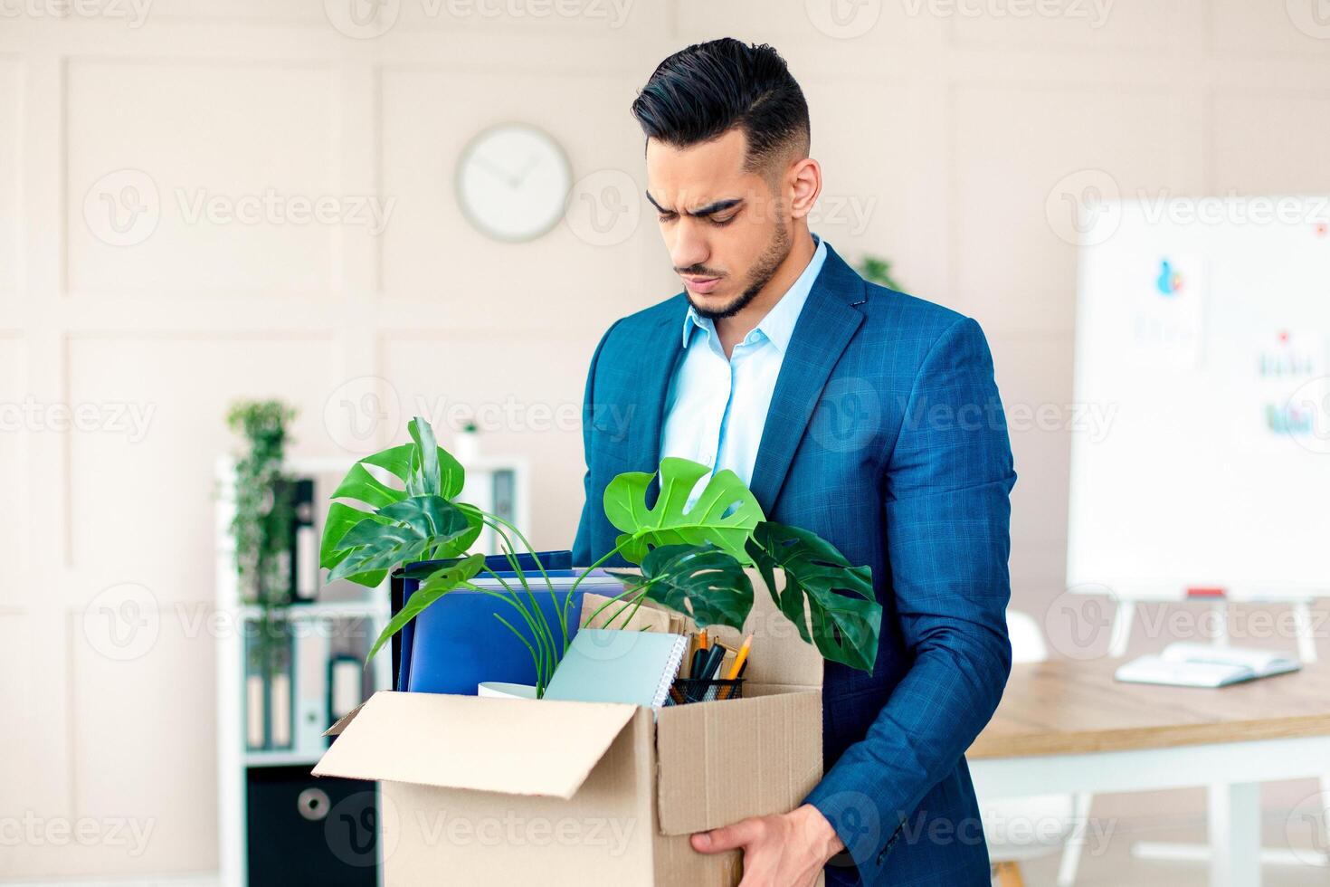 A young Arab entrepreneur stands in a modern office, holding a box filled with plants, files, and office supplies, expressing a serious demeanor as he prepares to leave. photo