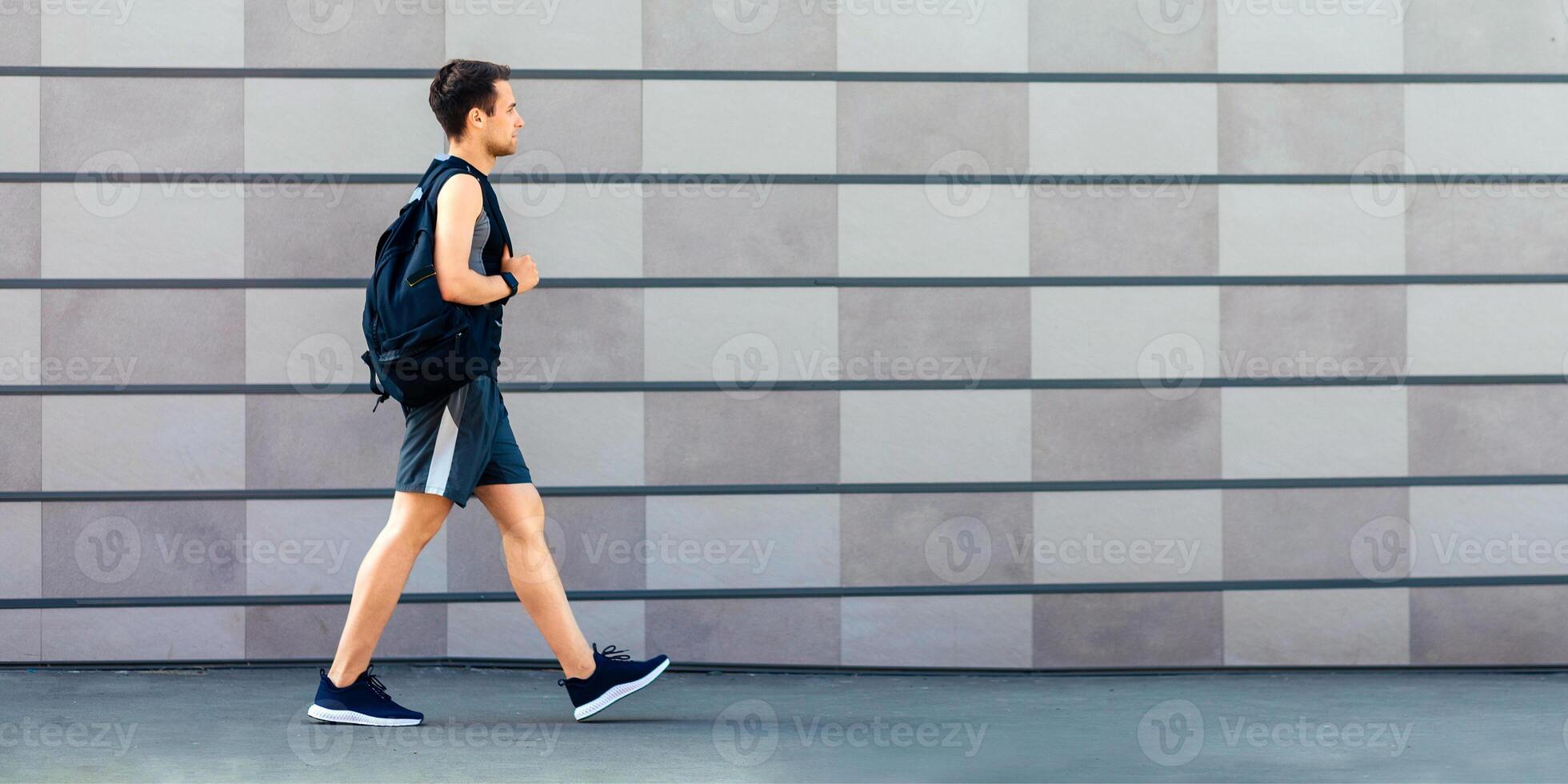 Go to morning workout. Guy in sportswear, sneakers and with backpack, on gray wall background, side view, free space photo