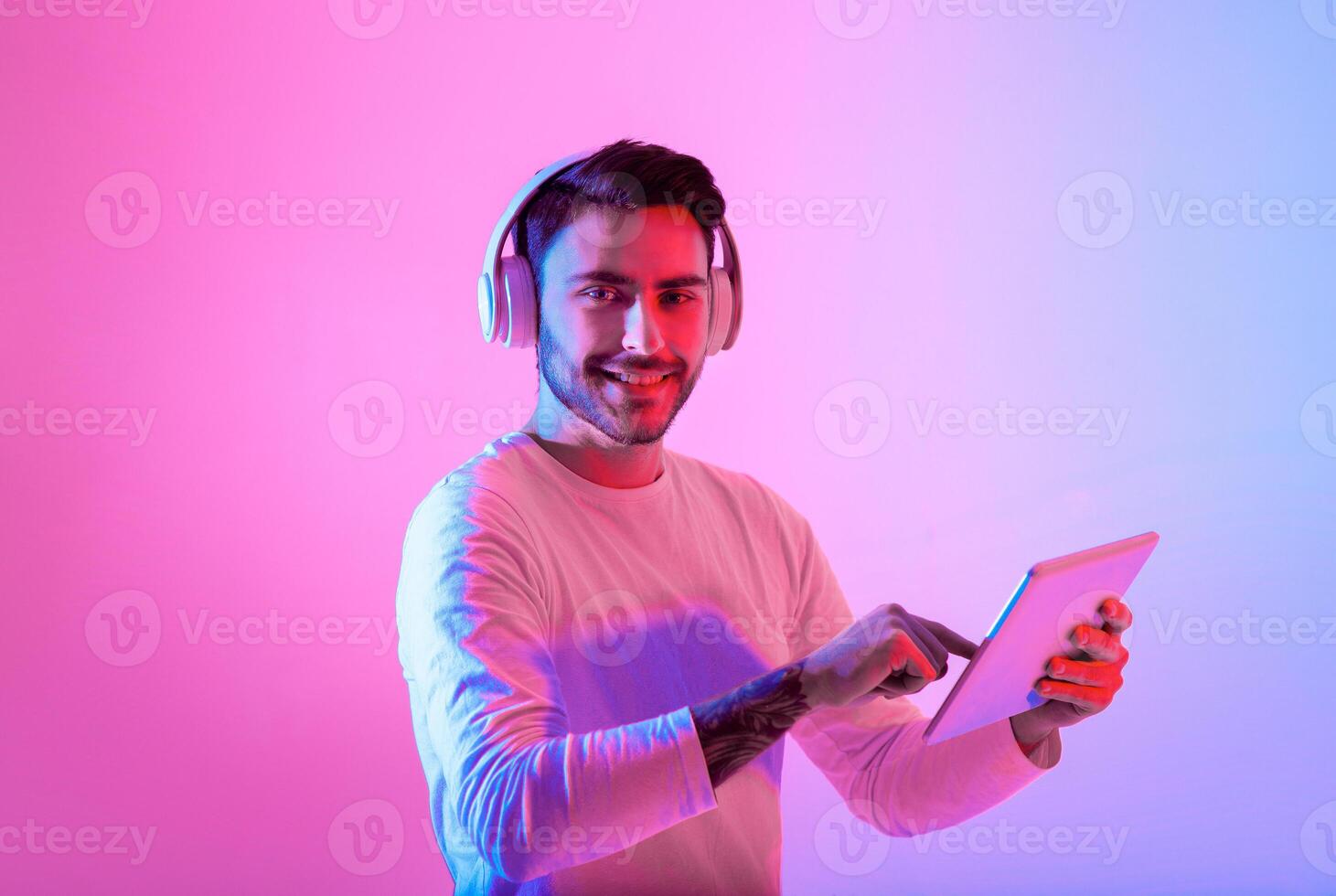 A young man smiles as he interacts with a tablet while wearing headphones. The background features bright pink and blue hues, creating a lively atmosphere. photo