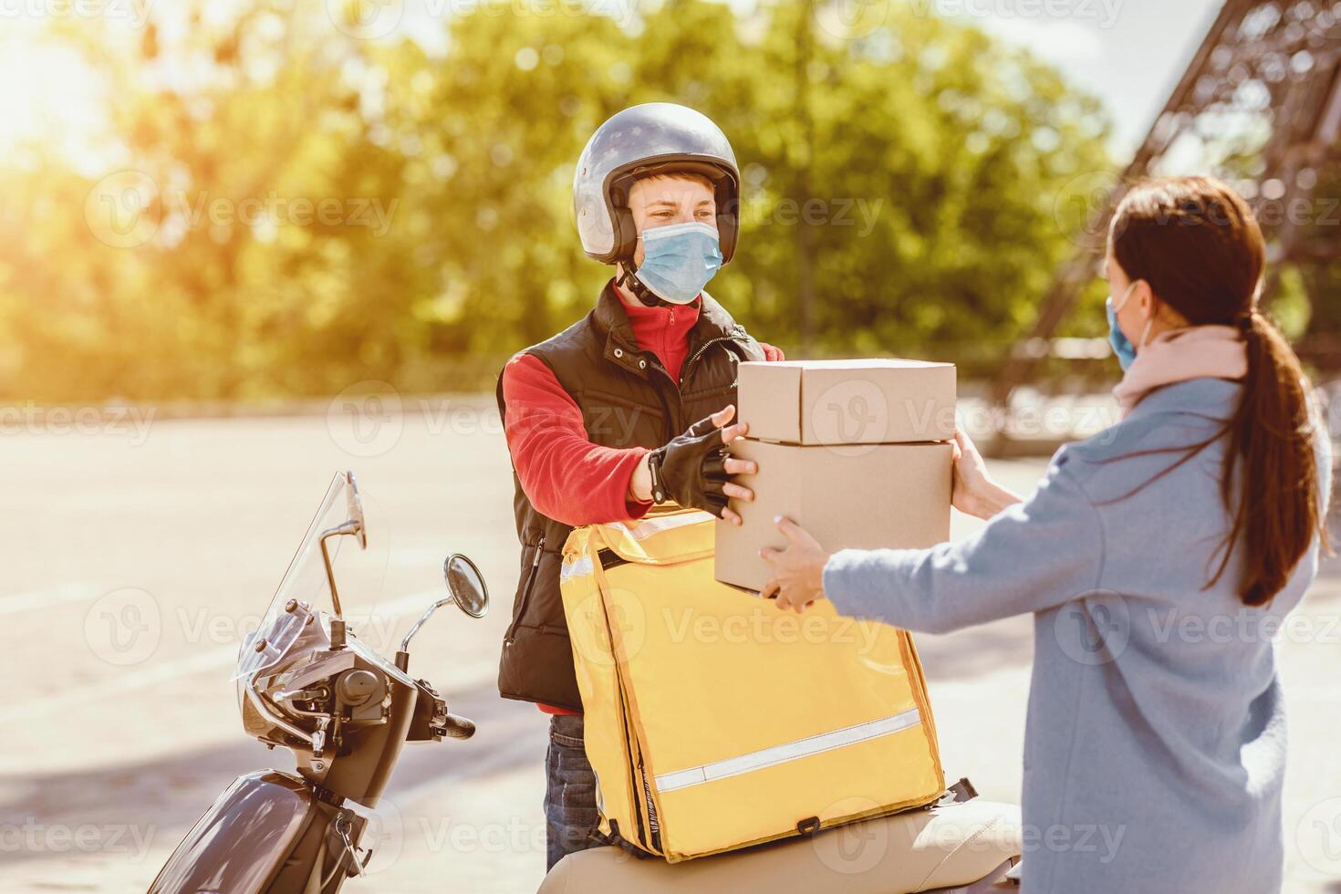 Safe Courier Service. Delivery Guy Giving Food Boxes To Customer Girl Standing Outdoors With Scooter, Wearing Medical Mask. photo