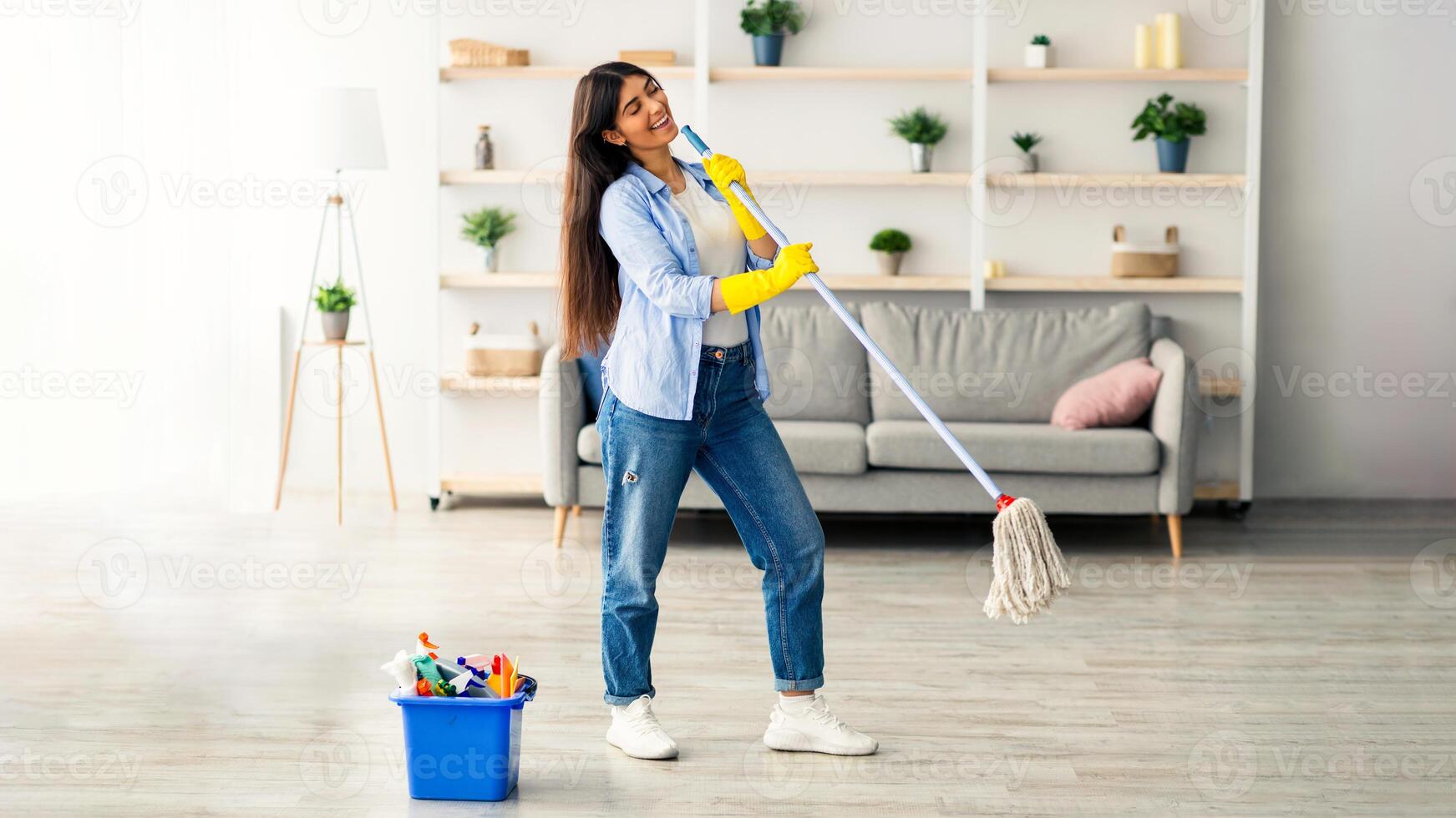 A young woman is happily cleaning her home using a mop. She is dressed casually and surrounded by a fresh and tidy living space, with plants adorning the shelves, copy space photo