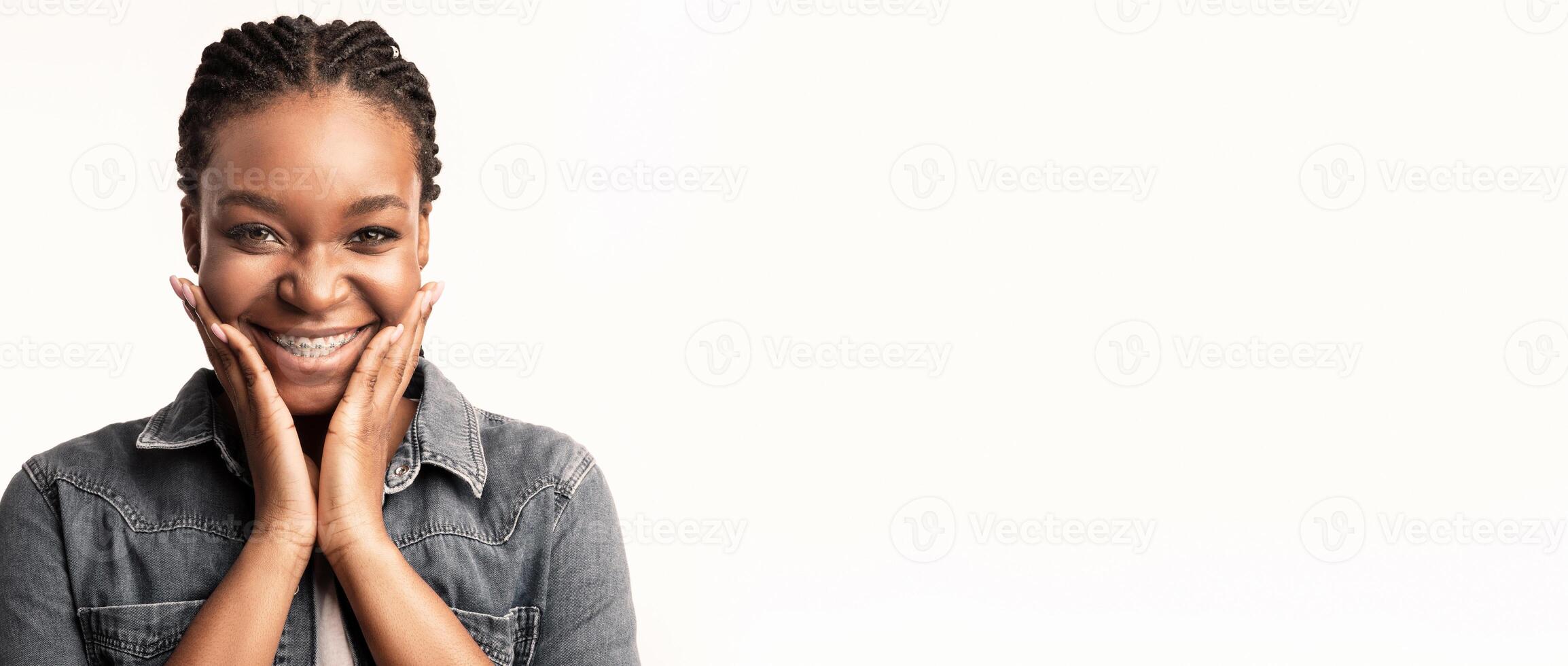 A woman stands with her hands on her cheeks, displaying a wide smile that radiates joy and positivity. She has braided hair and wears a denim jacket, exuding confidence and warmth. photo
