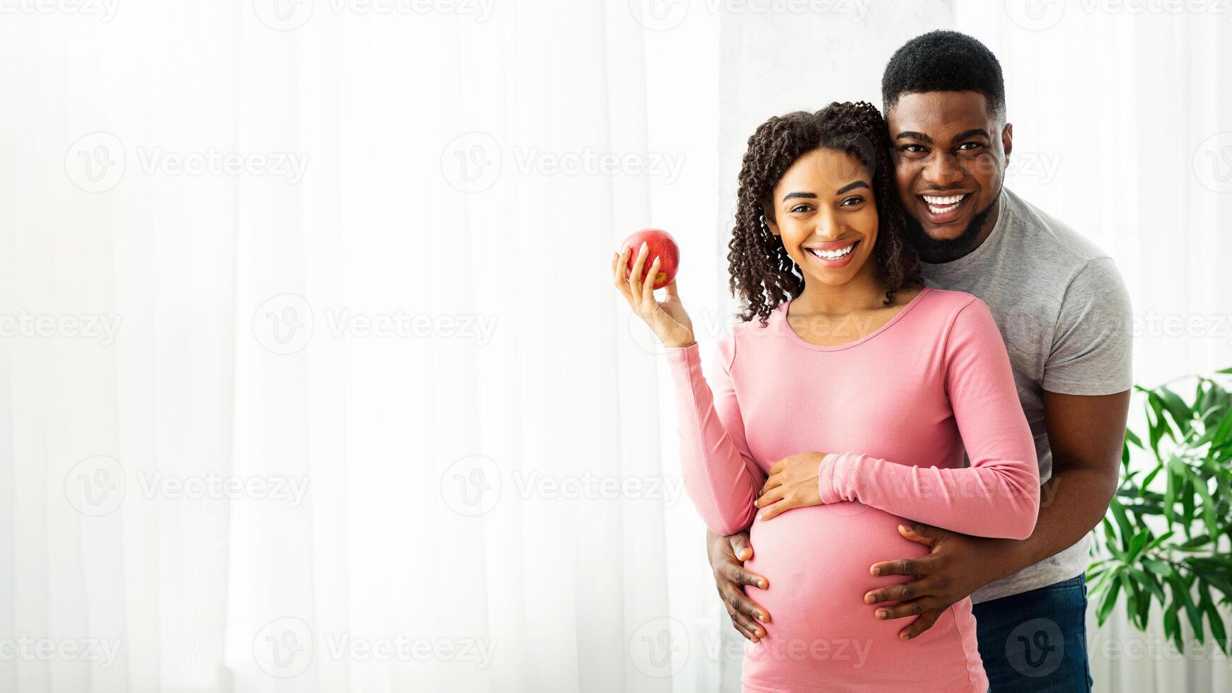 Cheerful african expecting couple posing at home next to window, lady holding fresh apple, copy space photo