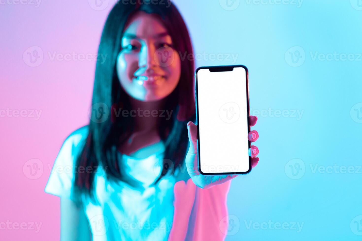 A young woman holds up a smartphone in front of her with a blank screen. The vibrant blue and pink background adds a modern touch to her confident pose in a studio setting. photo