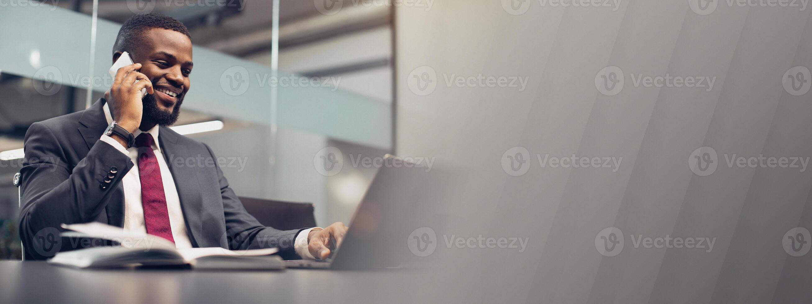 Successful young black businessman in expensive blue suit working on laptop at office, having phone conversation with his personal assistant or business partner and smiling, copy space photo