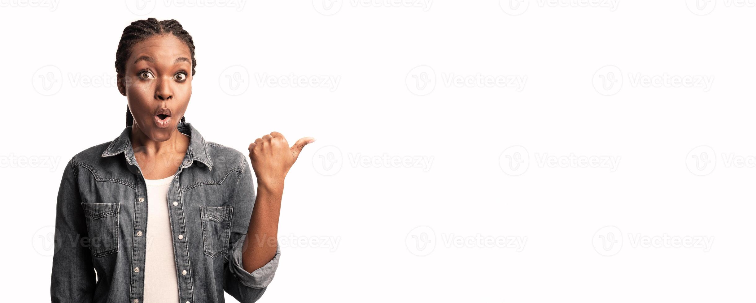 A young woman with braided hair expresses surprise while pointing to the right side, showcasing a moment of excitement or revelation in a clean, bright studio environment. photo