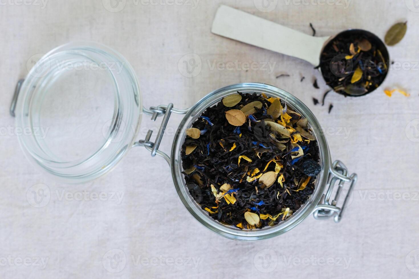A glass jar with loose leaf tea and metal spoon placed on a white linen cloth with tea leaves scattered around photo