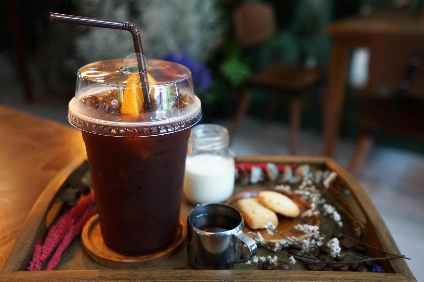 Plastic take away cup of iced black coffee served with milk, sugar syrup and butter cookies on wooden tray table decorated with dry flower and coffee beans photo