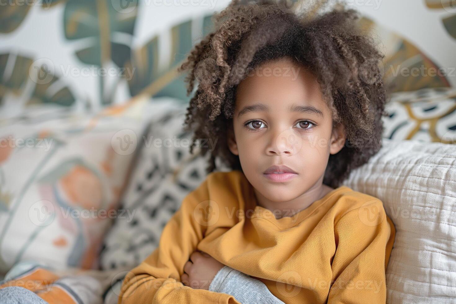 A young Black boy with curly hair looks thoughtfully at the camera, showcasing a cozy indoor setting with patterned cushions. photo