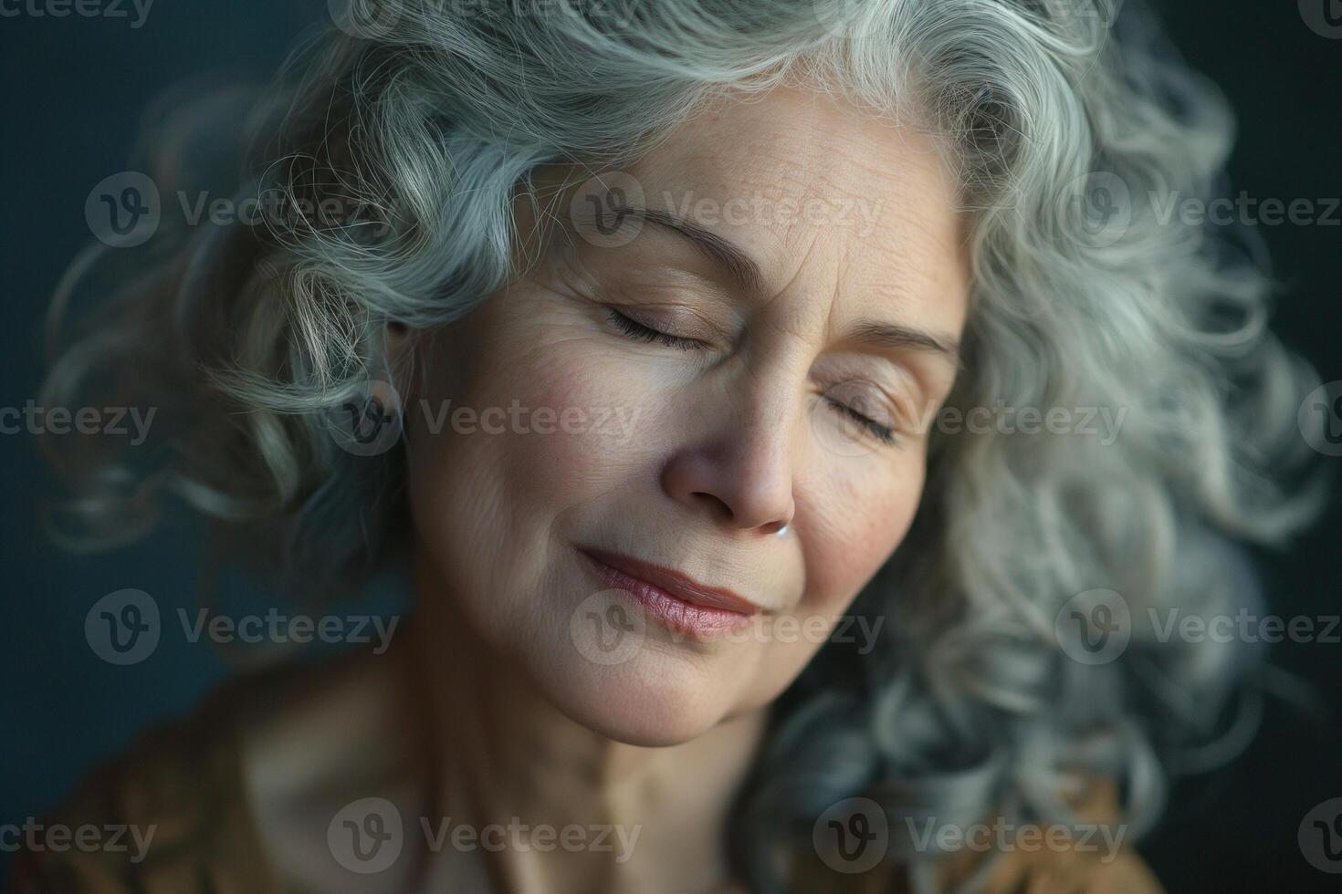 A serene older woman with silver curly hair closes her eyes, exuding calmness and wisdom against a soft, blurred background. photo