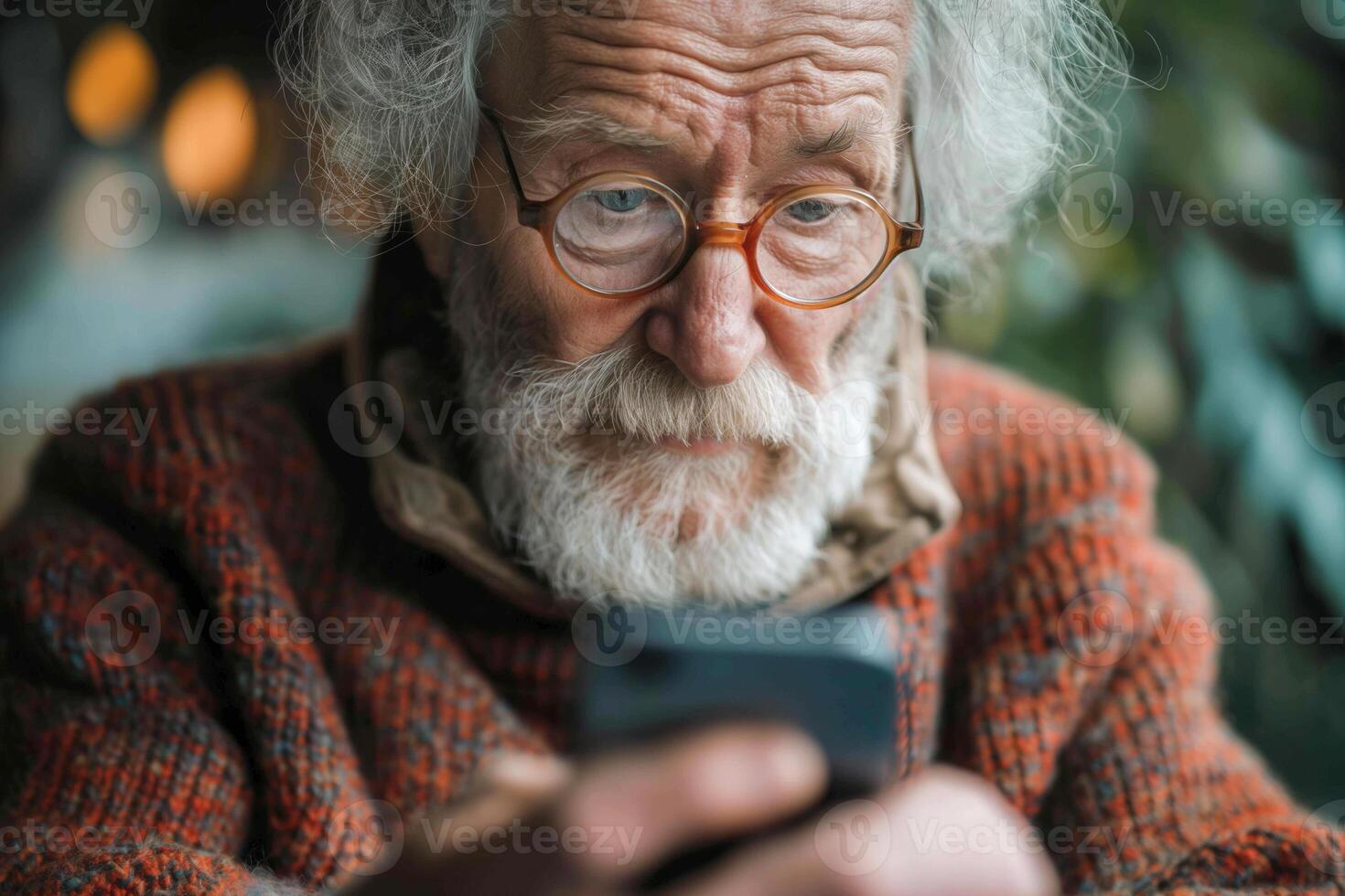 An elderly Caucasian man with a beard and glasses engrossed in his smartphone, sitting indoors surrounded by greenery. photo