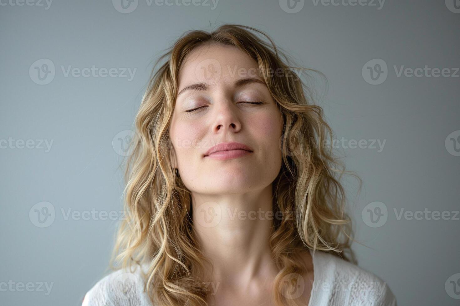 A serene young Caucasian woman with curly hair, eyes closed, reflecting tranquility against a soft gray background. photo