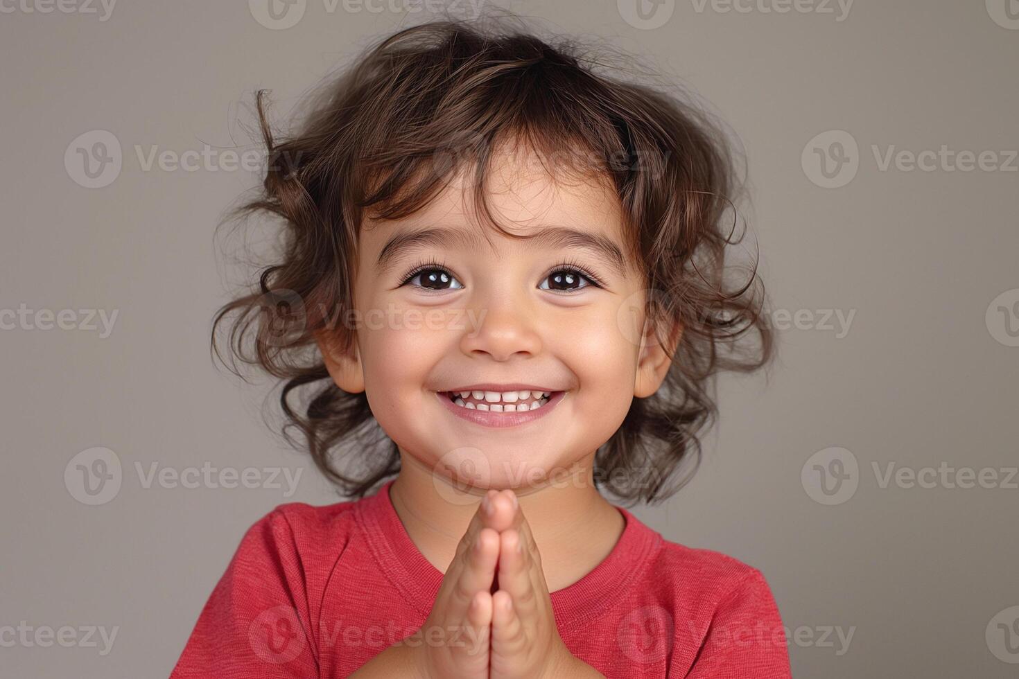 A joyful young boy of Hispanic descent with curly hair expresses happiness, clasping his hands together in a playful manner. photo