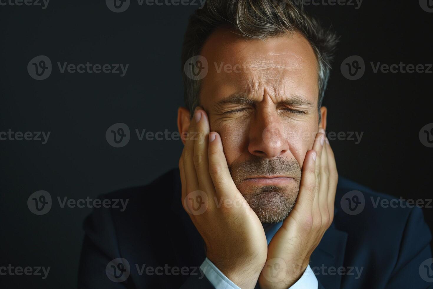 A stressed middle-aged Caucasian man in a dark suit expresses frustration while resting his head in his hands against a dark background. photo