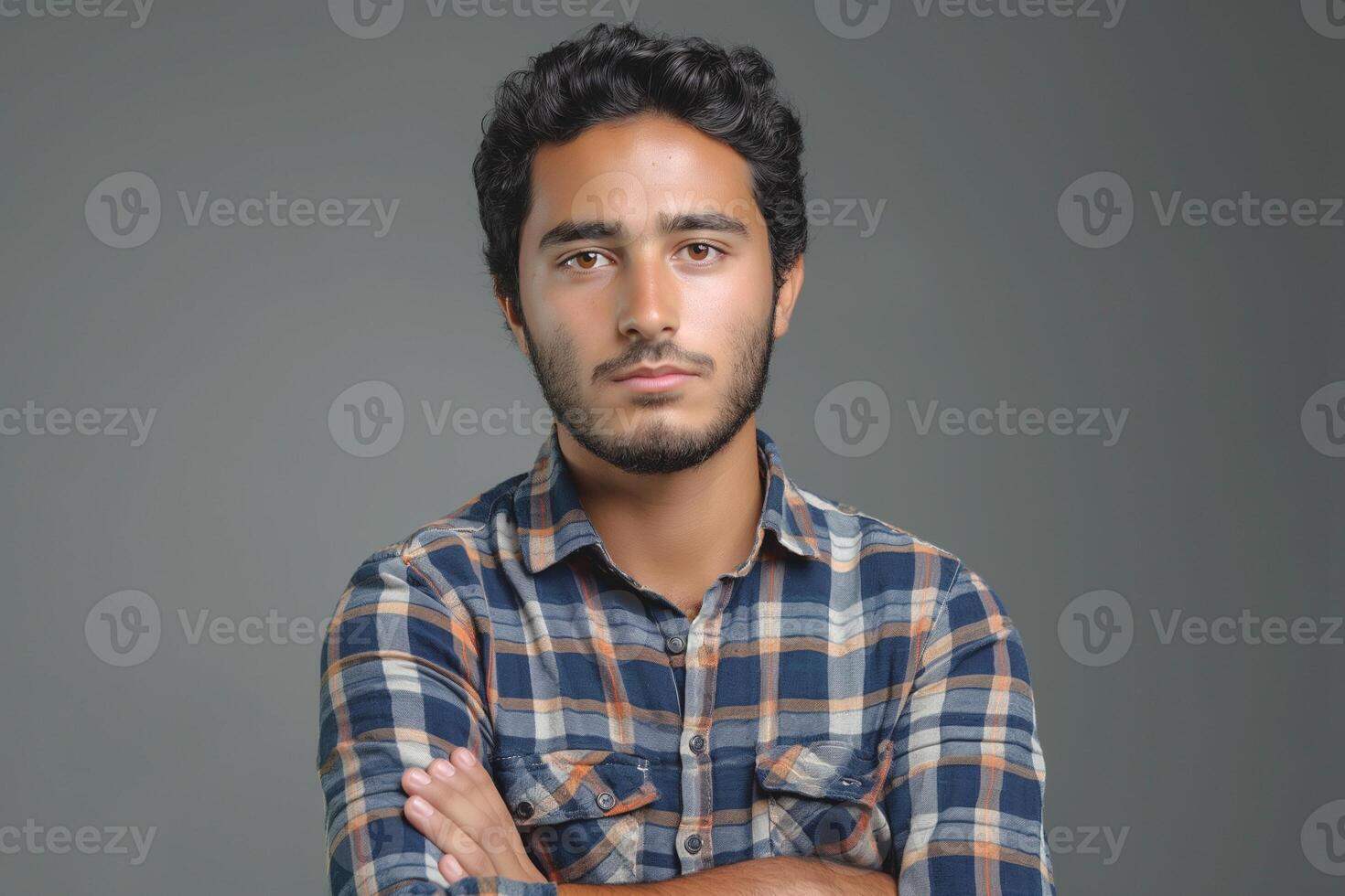 Young South Asian man with dark curly hair, wearing a plaid shirt, presents a confident pose against a neutral background. photo
