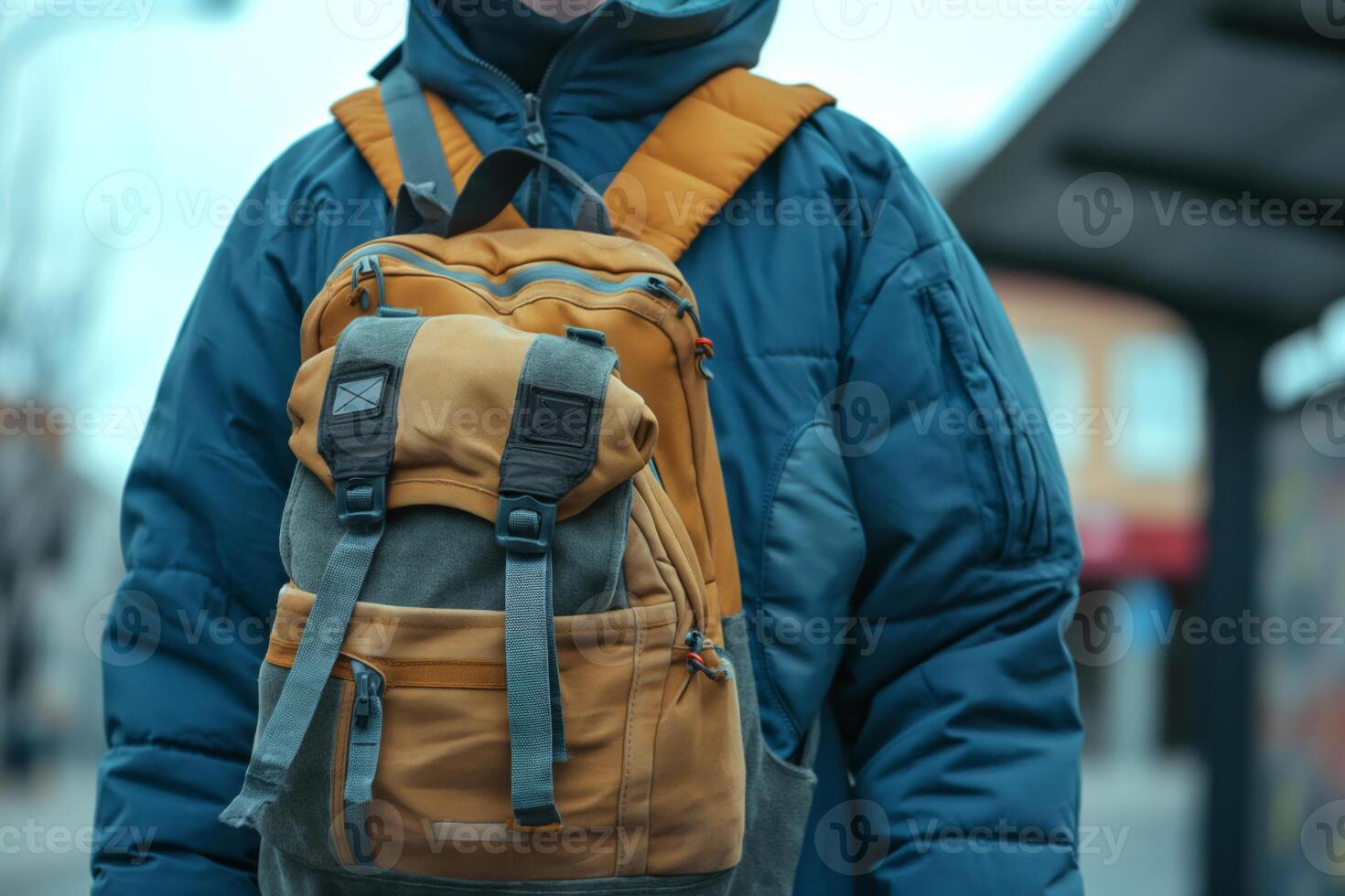 A young male wearing a warm blue coat and a stylish mustard backpack, standing at a bus stop in a chilly urban setting. photo