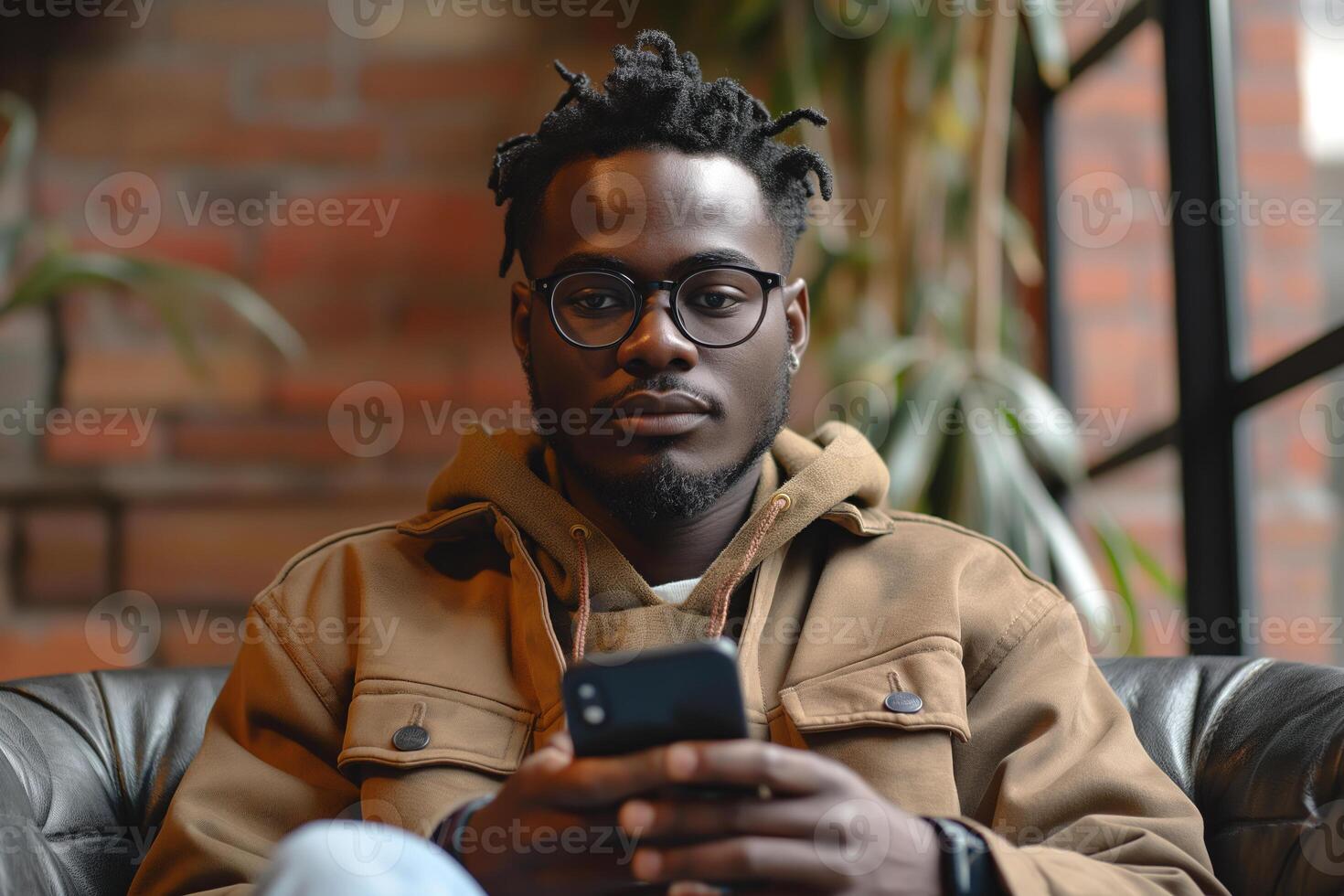 A young Black man with glasses sits comfortably indoors, focused on his smartphone amidst a cozy atmosphere. photo