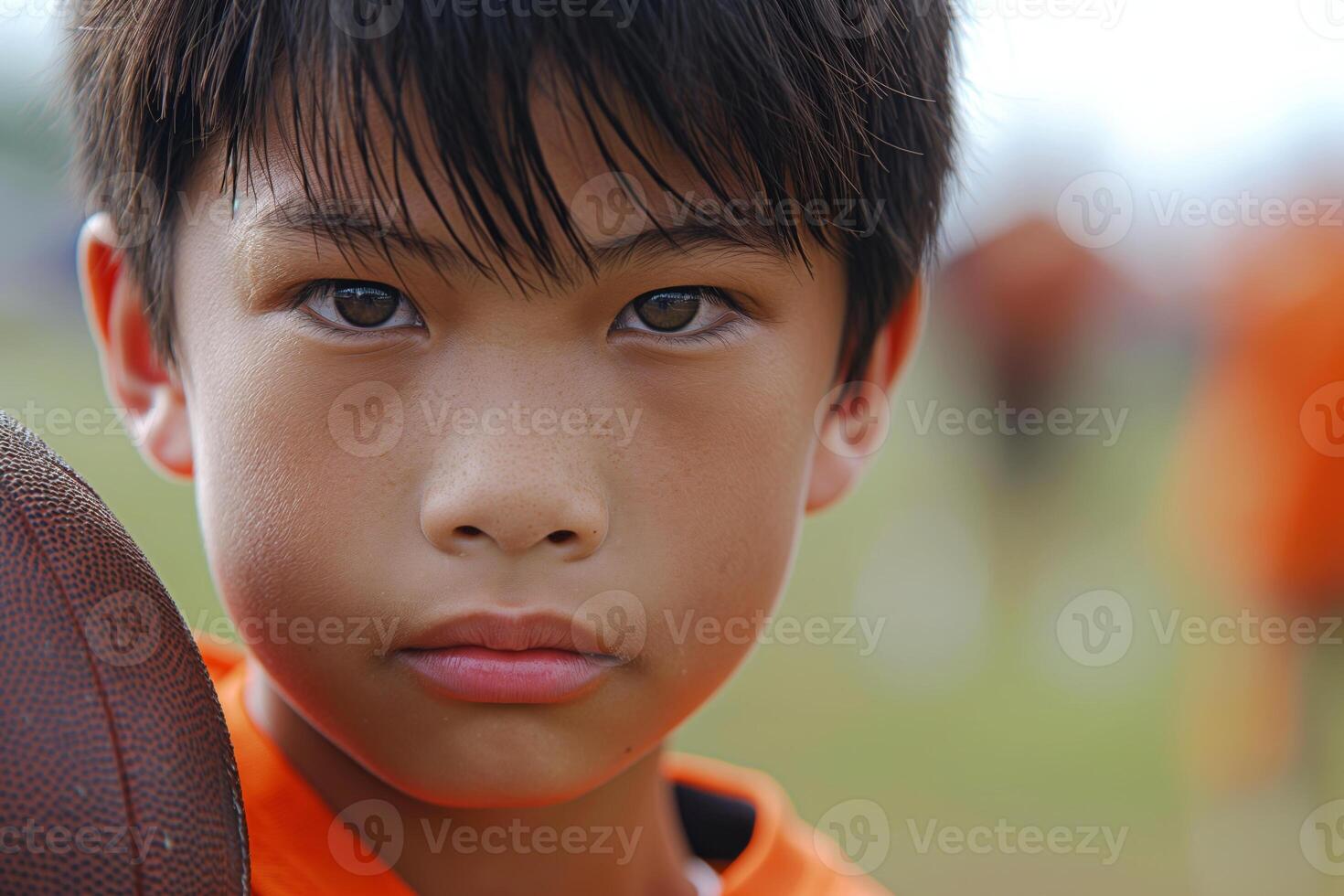 Close-up portrait of a young Asian boy with a determined expression, holding a football. He wears an orange jersey. photo