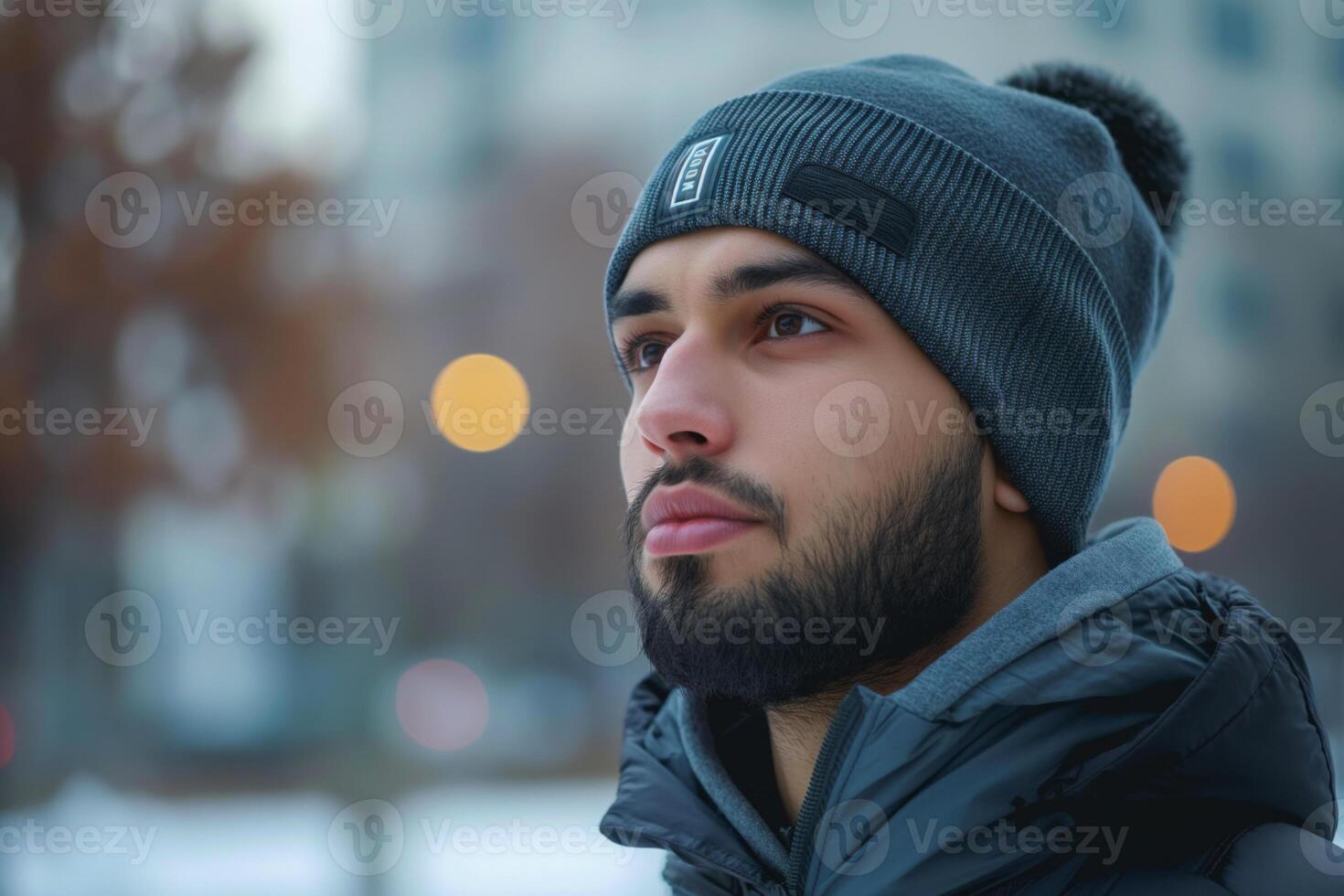 A young Middle-Eastern man gazes thoughtfully while wearing a gray beanie and a dark jacket in a cold urban setting. photo