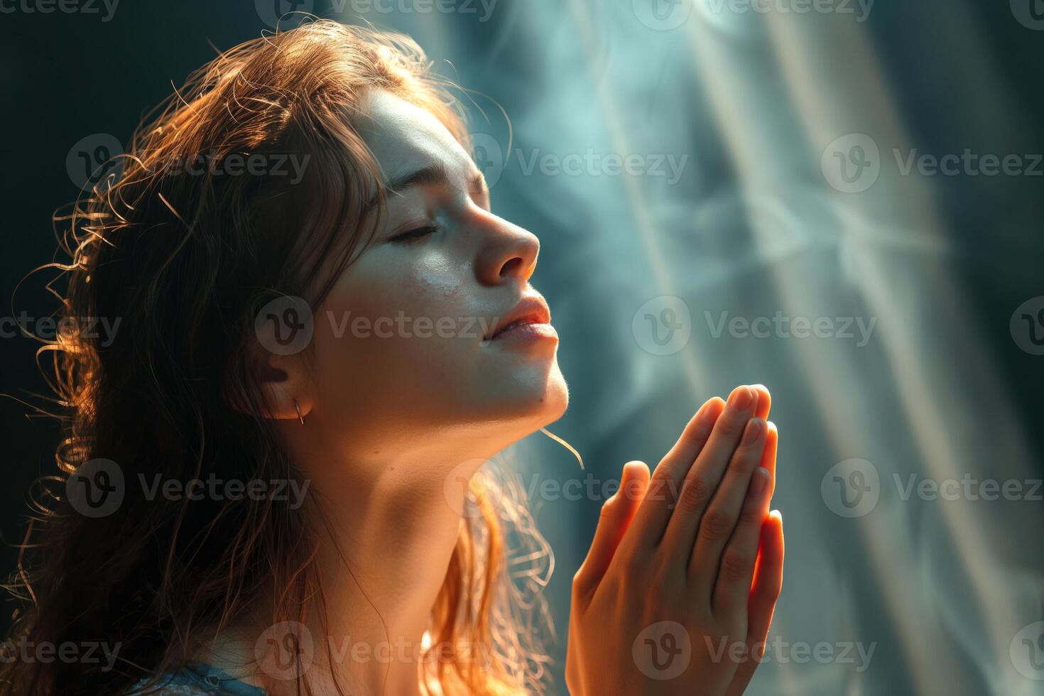 A young Caucasian woman with closed eyes and hands clasped in prayer, illuminated by soft, ethereal light and gentle smoke. photo