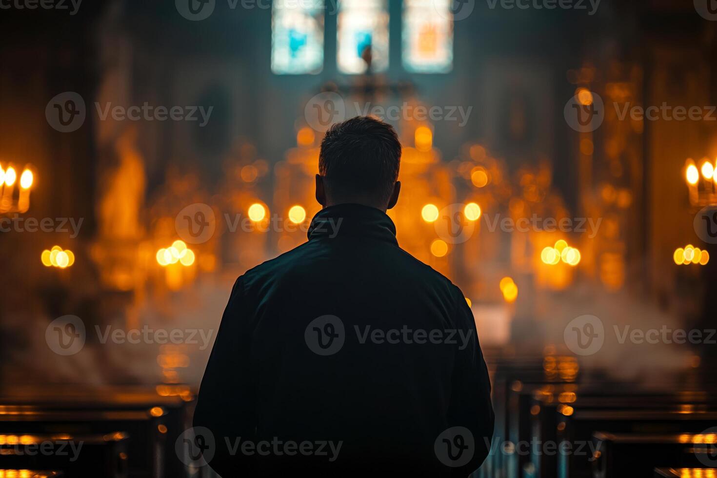 A contemplative man in a dark jacket stands in an illuminated church, surrounded by soft candlelight and ornate decor. photo
