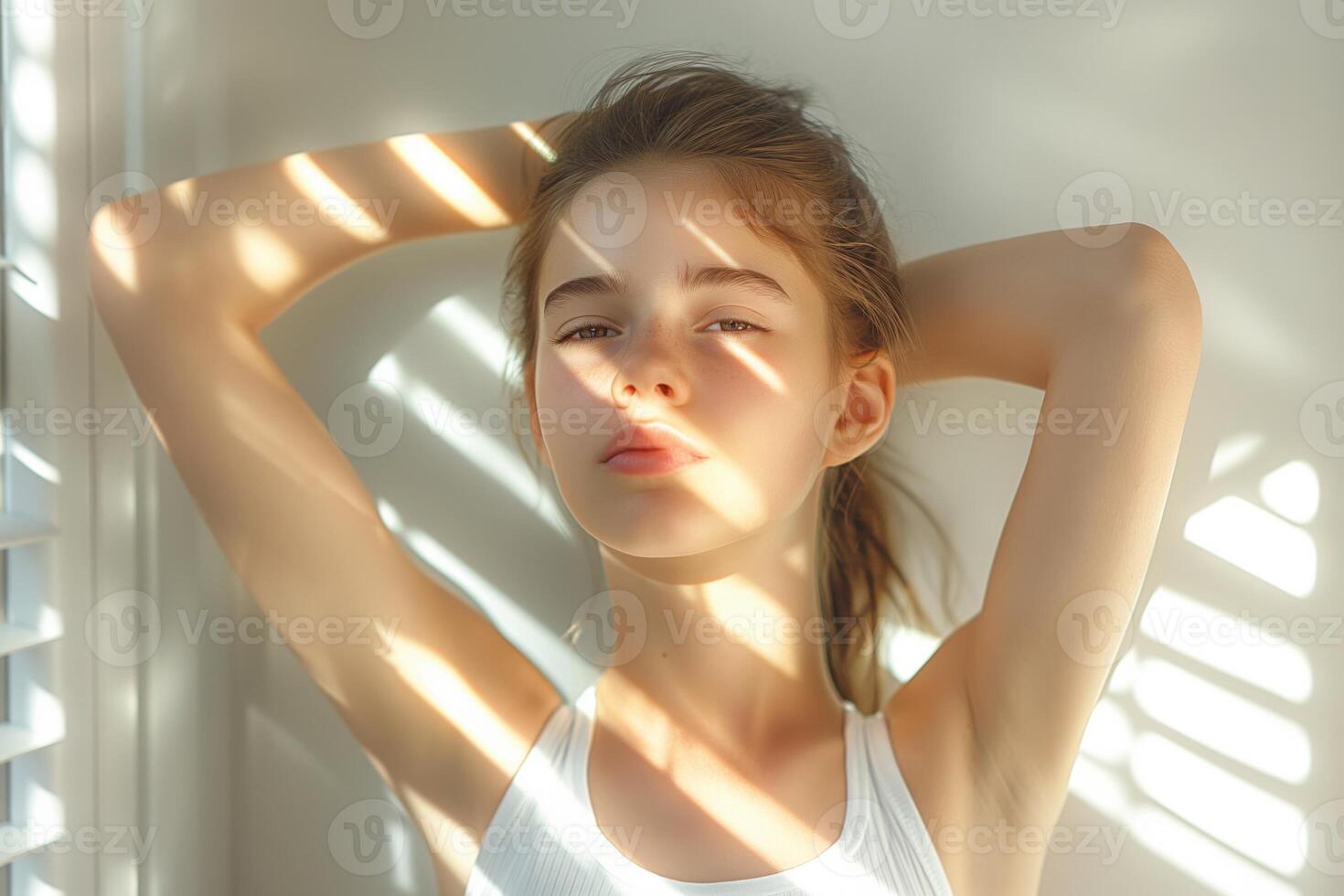 A young Caucasian girl in a white tank top poses against a sunlit wall, with shadows creating a striking pattern across her face. photo