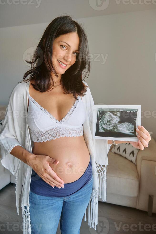 Smiling pregnant woman holding sonogram in living room photo