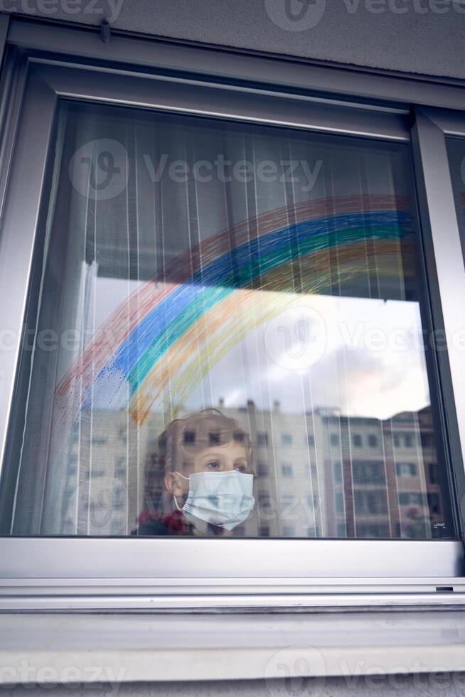 Boy in mask standing near window with rainbow at home photo