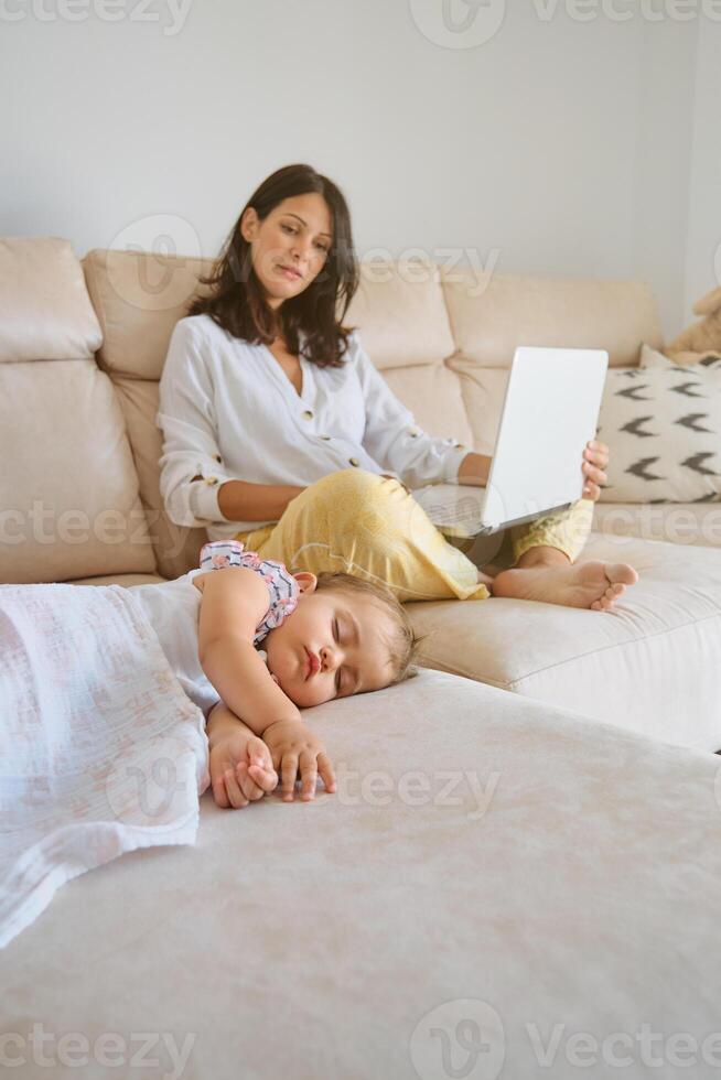 Woman browsing netbook while sitting on sofa near napping baby photo
