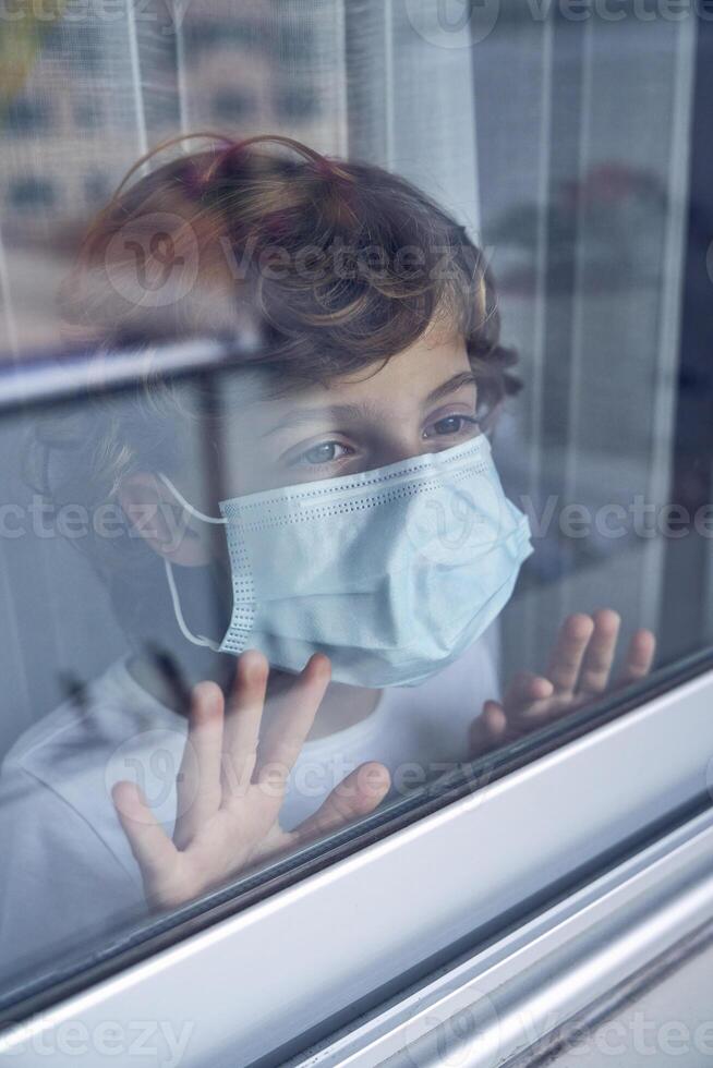 Pensive boy in mask standing alone and looking out of window at photo
