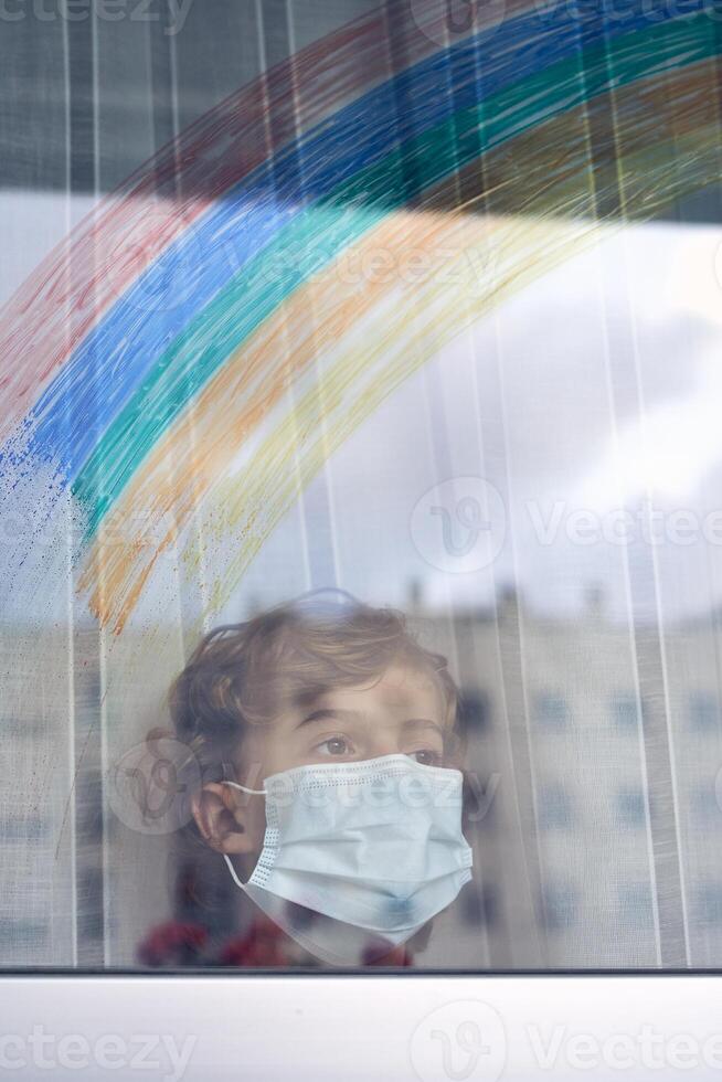 Boy with face mask standing near window with reflection of rainb photo