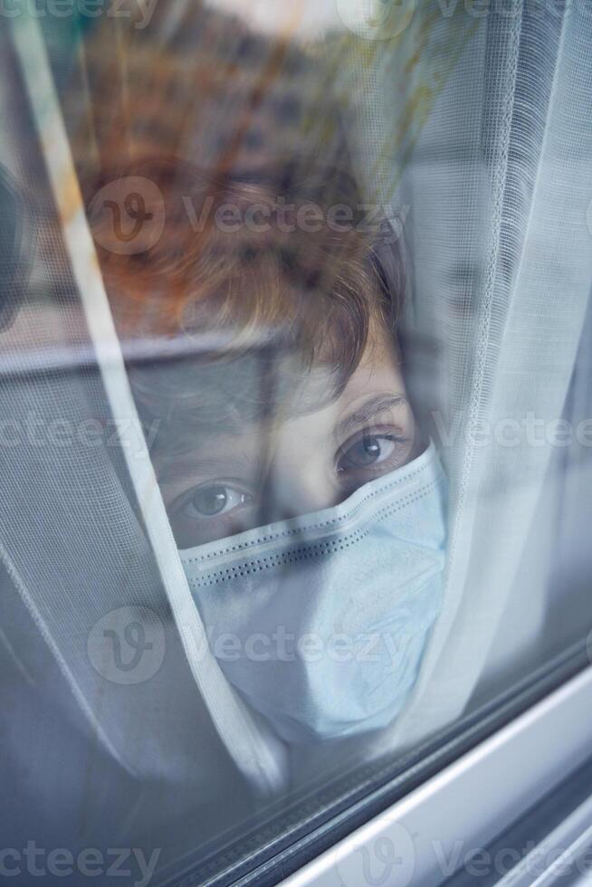 Cute boy in mask looking through window at home photo