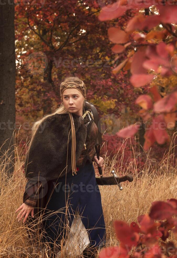 warrior girls with sword in hands in autumn forest. blonde dressed in historical costume of warrior woman, with brown fur cloak, medieval dress. warrior girl holds steel sword in hands, foreground photo