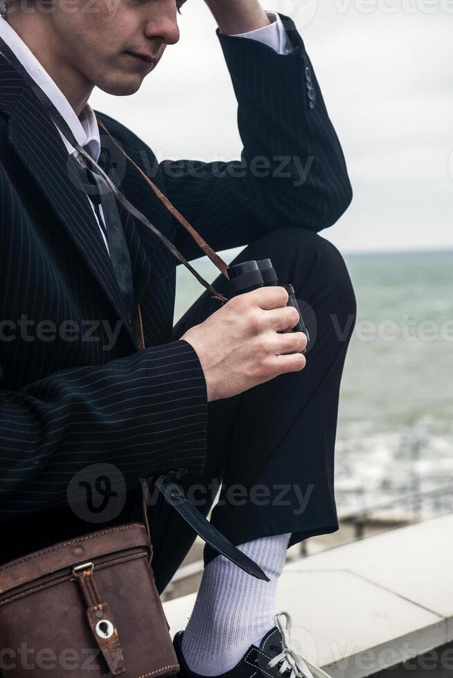 Portrait of a young man against the backdrop of the sea, he is looking into the distance through binoculars. He has long hair, is wearing a suit and tie. Relaxed, peaceful, thoughtful, happy and free on the seashore photo