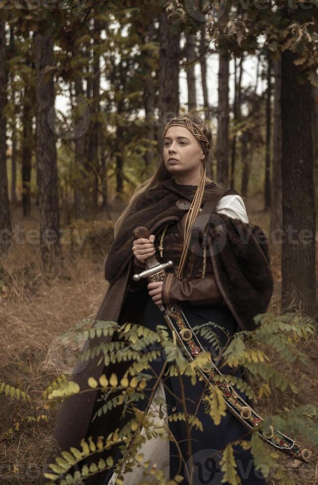 warrior girls with sword in hands in autumn forest. blonde dressed in historical costume of warrior woman, with brown fur cloak, medieval dress. warrior girl holds steel sword in hands. photo