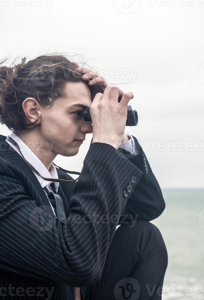Portrait of a young man against the backdrop of the sea, he is looking into the distance through binoculars. He has long hair, is wearing a suit and tie. Relaxed, peaceful, thoughtful, happy and free on the seashore. photo