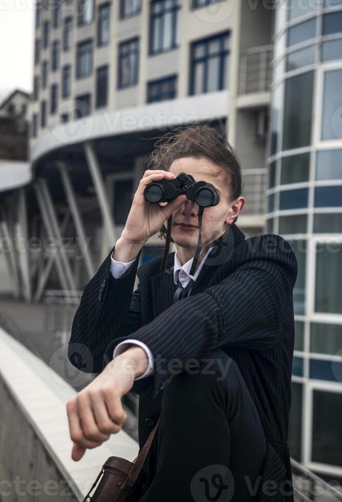 Portrait of a young man against the backdrop of modern architecture, he is looking through binoculars. He has long hair, he is wearing a suit. Front view, close-up. Relaxed, peaceful, thoughtful, happy photo