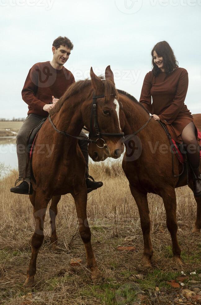 Young romantic couple horse riding, against the background of a field with autumn grass and a lake. They gallop across the fields, stylishly dressed in brown clothes. They smile photo