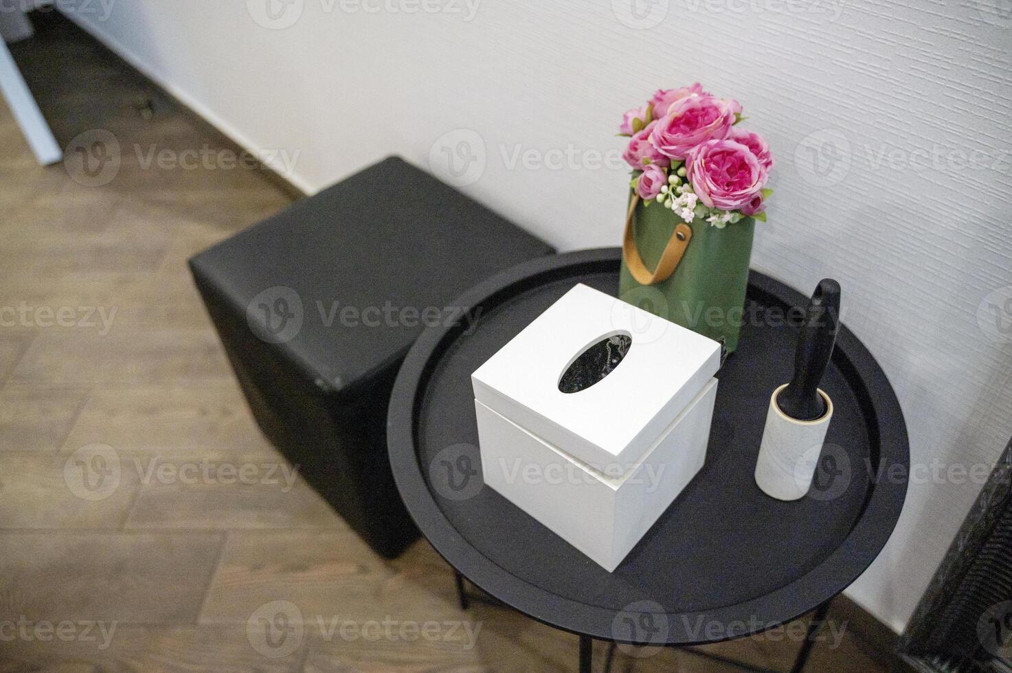 round black metal table next to it pouf. on the table are a box for napkins, decorative flowers, a brush for cleaning clothes. top view. on the floor are wood-like tiles. the walls are white. photo