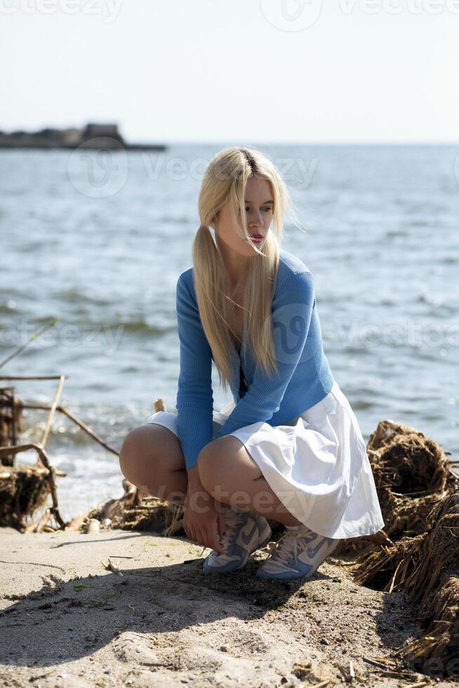 beautiful blonde girl sits on a sandy beach in sunny weather. girl in a short white skirt and blue blouse draws a heart on the sand with her hand. against the background of a calm sea. photo