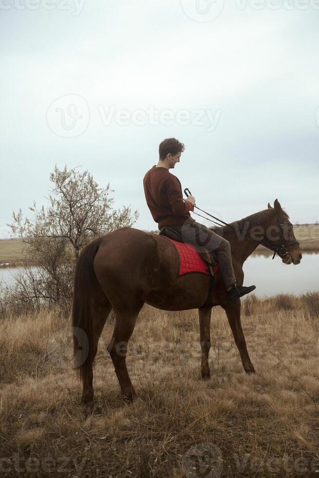 A man in a brown sweater rides uphill on a horse across a field. Around him are fields with autumn grass. A lake is visible on the horizon. Concept of horse riding outdoors, sport and recreation. A man riding a horse in a field photo