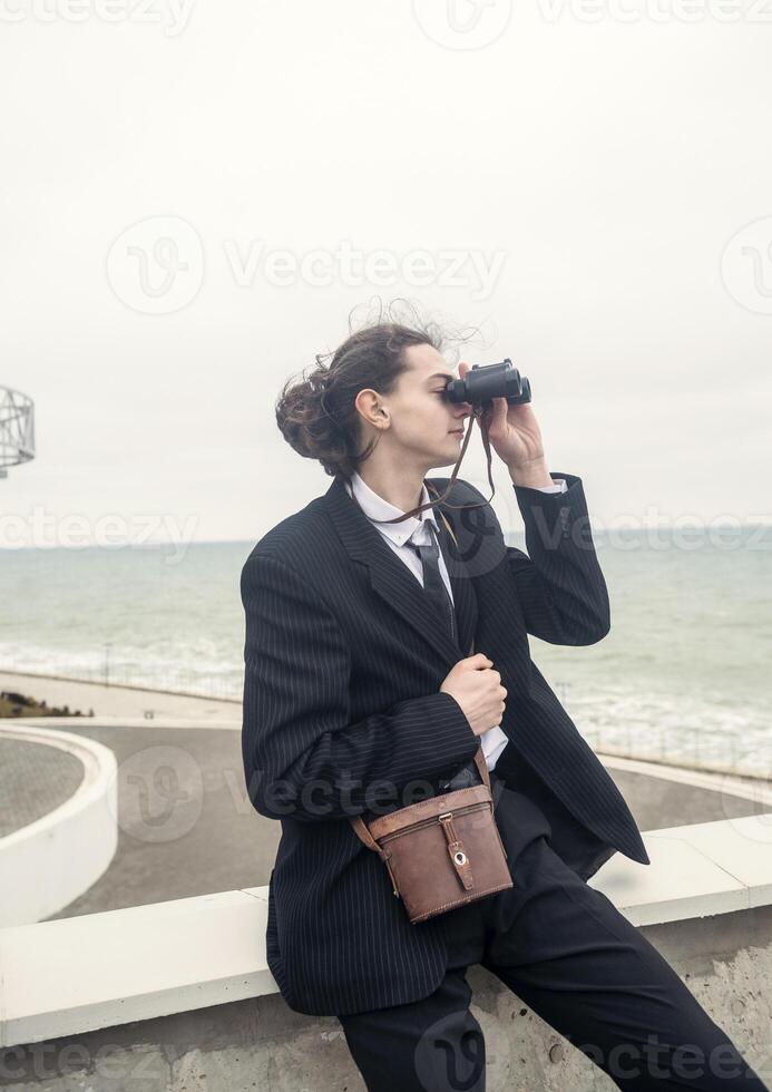 A young man sits alone against the backdrop of modern architecture near the sea. He looks through binoculars at the sea. He has long hair, wears a suit and tie. Relaxed, peaceful, thoughtful photo