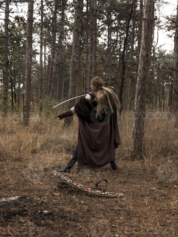 warrior girl with sword in hands in the forest. blonde dressed in historical costume of female warrior, trains with sword in hands. dressed in long cloak. warrior raised sword, attacks enemy photo