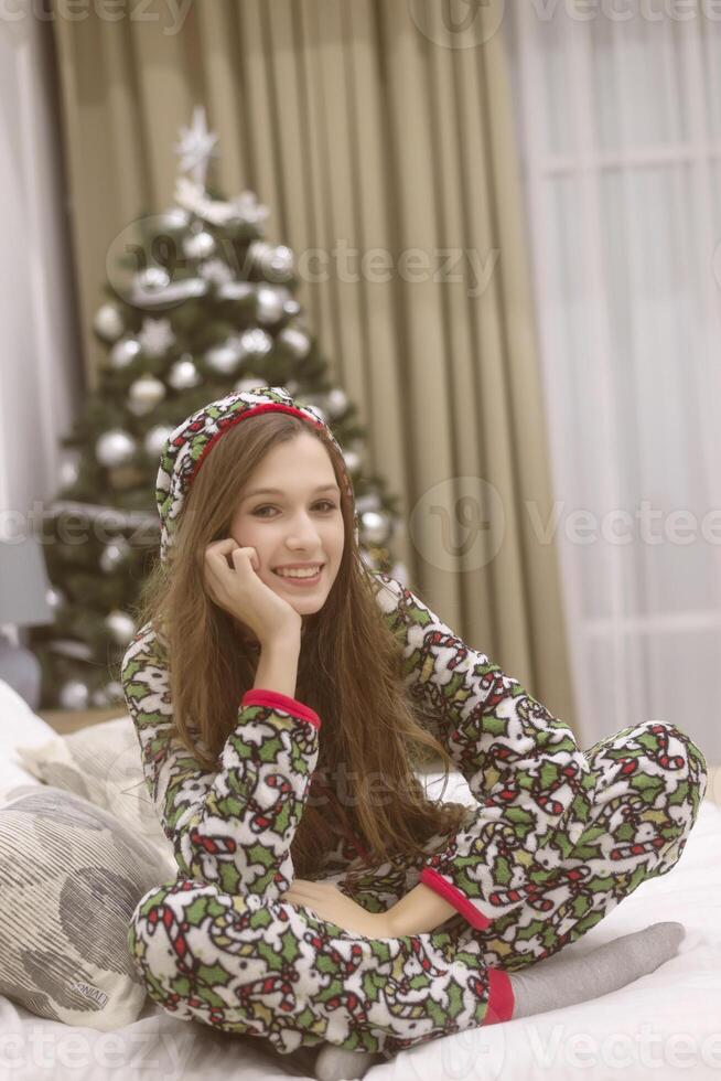 Beautiful young woman in pajamas sits on the bed against the background of a Christmas tree. She smiles and makes wishes. Christmas and New Year concept. Looking at the camera photo