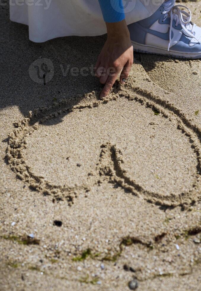 Heart sign drawing on the yellow beach sand. High quality photo. A woman's hand on the beach drew a heart sign on the sand. You can see a woman's hand, sneakers and a girl's white skirt. photo