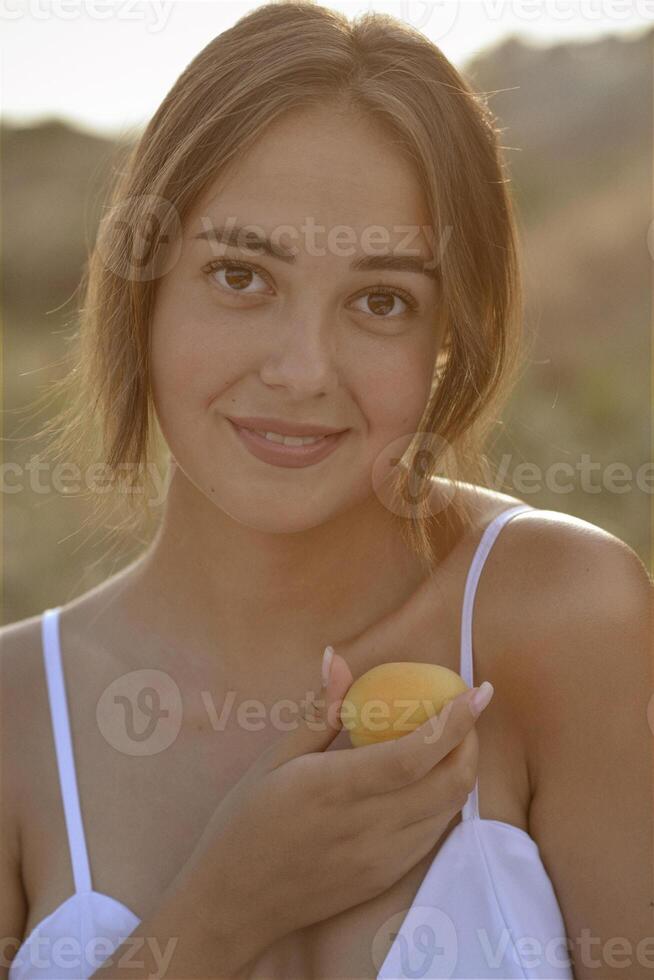 close-up of a beautiful young girl with a yellow apricot in her hands near her face. she smiles sweetly. posing with bare shoulders outdoors in the rays of the setting sun. concept of vitamins, summer holidays, cosmetics, self-care photo
