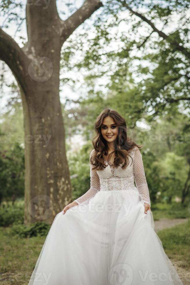 young girl bride in a white dress in a spring forest on a wedding day. beautiful brunette in a long stylish plan among huge trees. girl smiles and looks at the camera photo