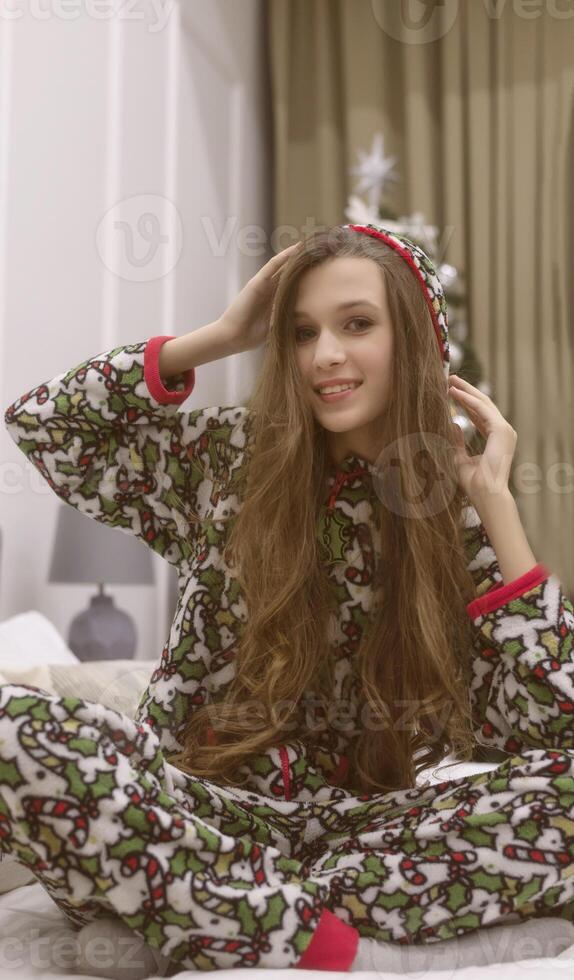 Beautiful young woman in pajamas sits on the bed against the background of a Christmas tree. She smiles and makes wishes. Christmas and New Year concept. Looking at the camera. photo