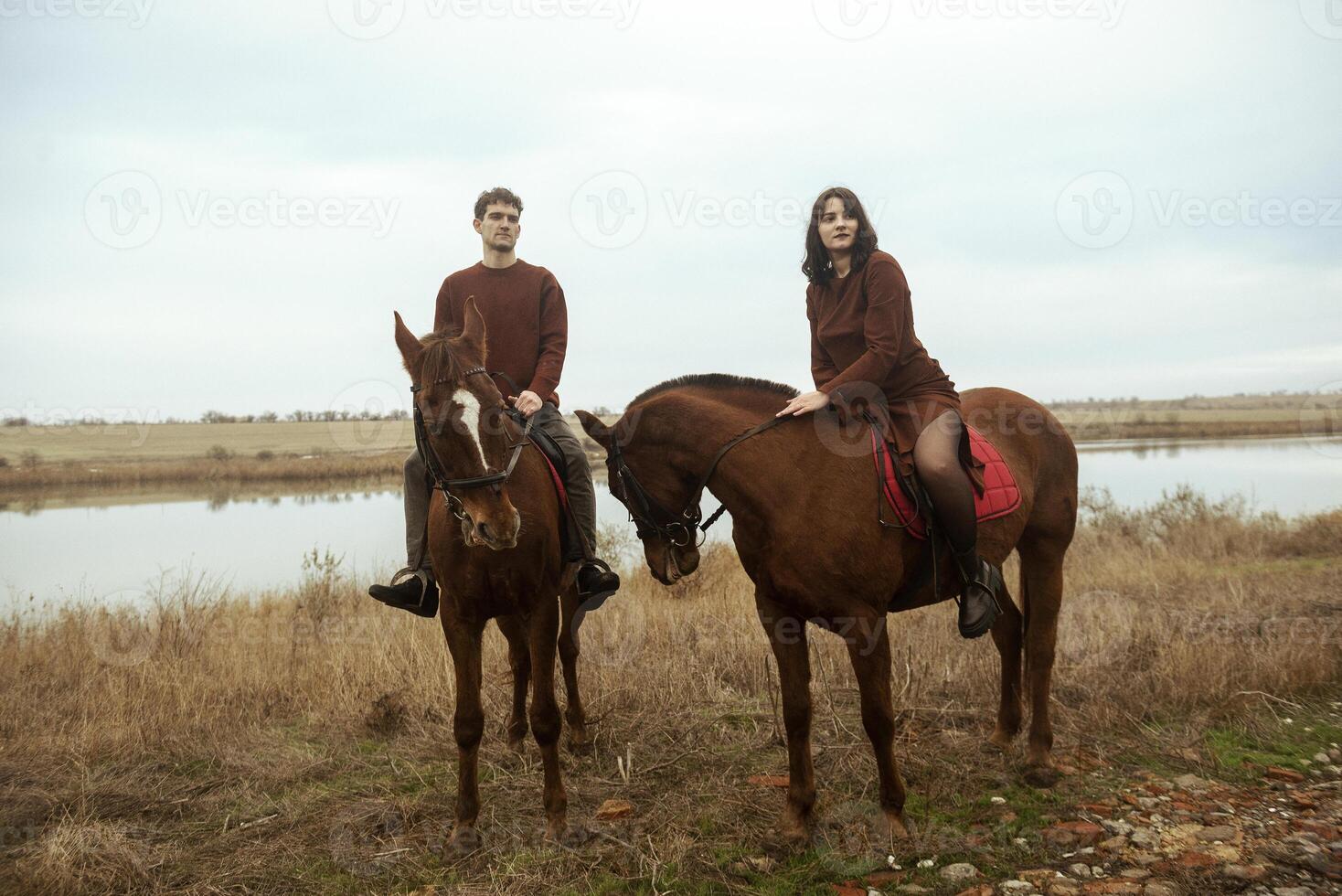Young romantic couple horse riding, against the background of a field with autumn grass and a lake. They gallop across the fields, stylishly dressed in brown clothes. They are happy during a romantic honeymoon. photo
